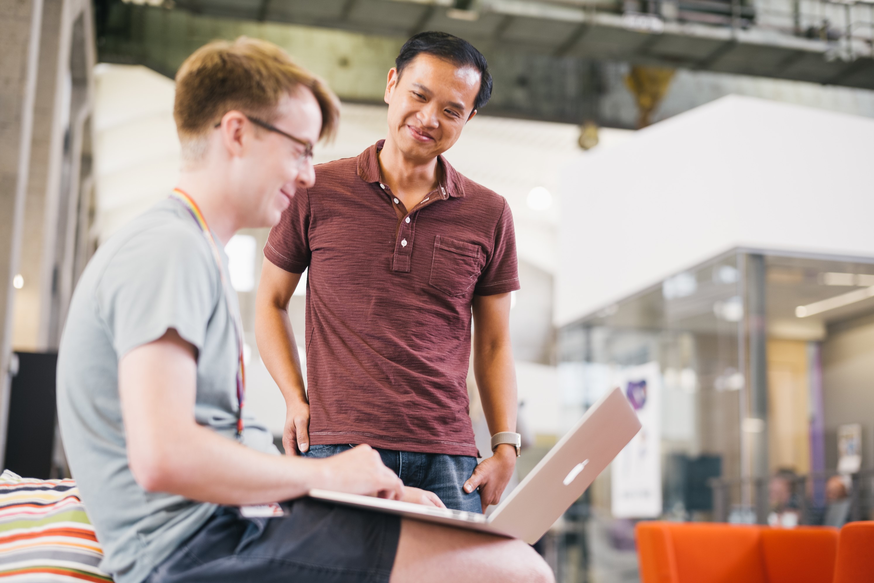 Two smiling men, one siting with a laptop on his lap  and the other standing, talking